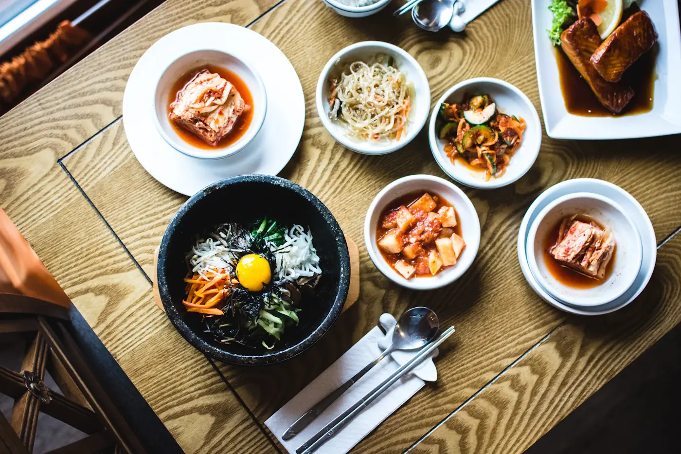 A wooden table set with bibimbap in a stone bowl and small dishes of kimchi, sprouts and braised fish