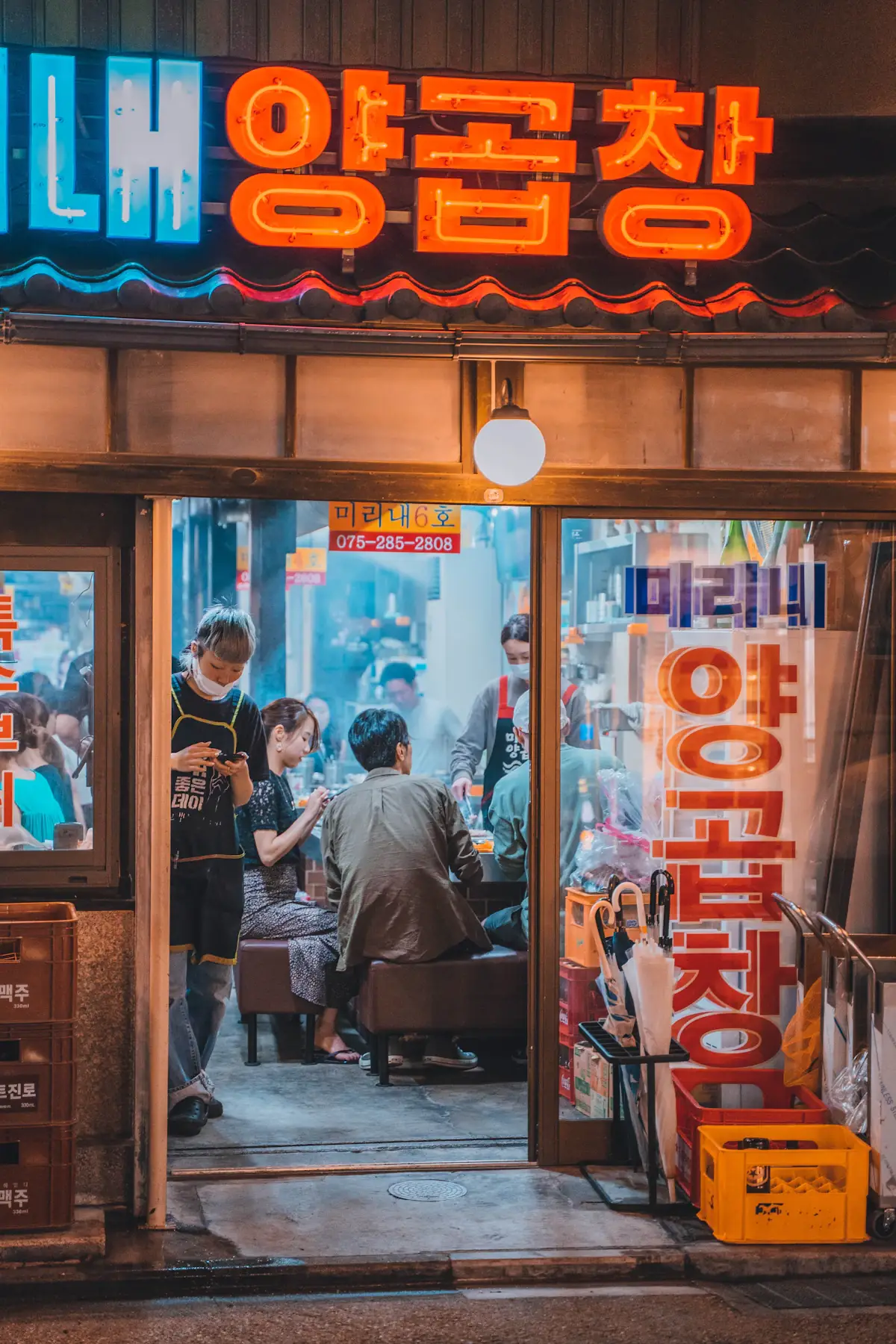 People eating inside a small Korean restaurant at night, seen through its window under red Hangul signs