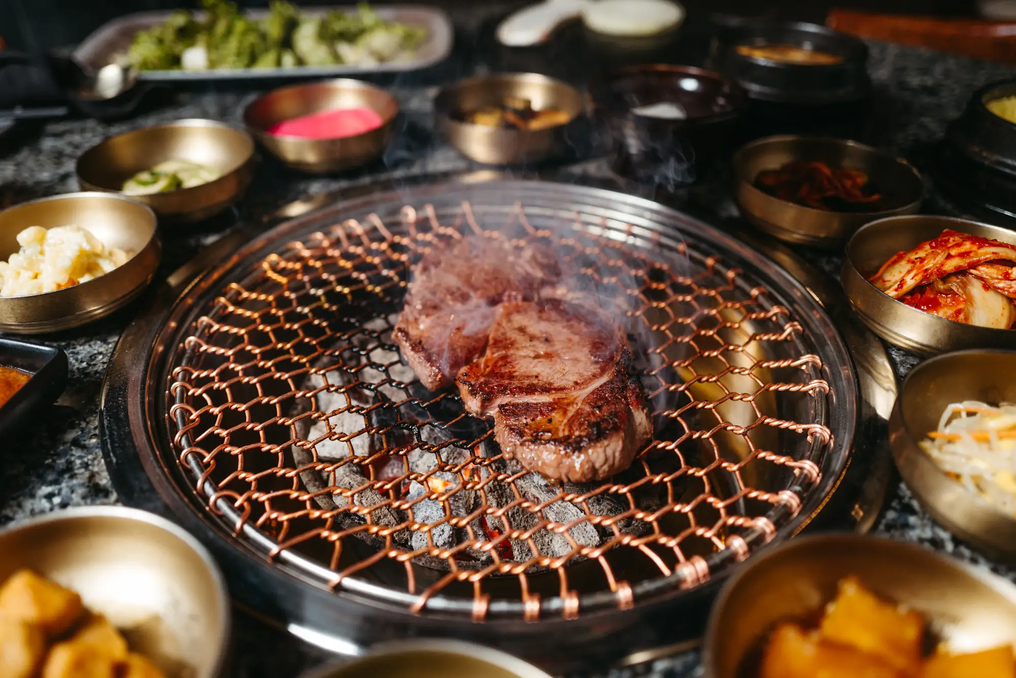 A round charcoal grill seen from above, sliced meat on the grate and small dishes of banchan all around it
