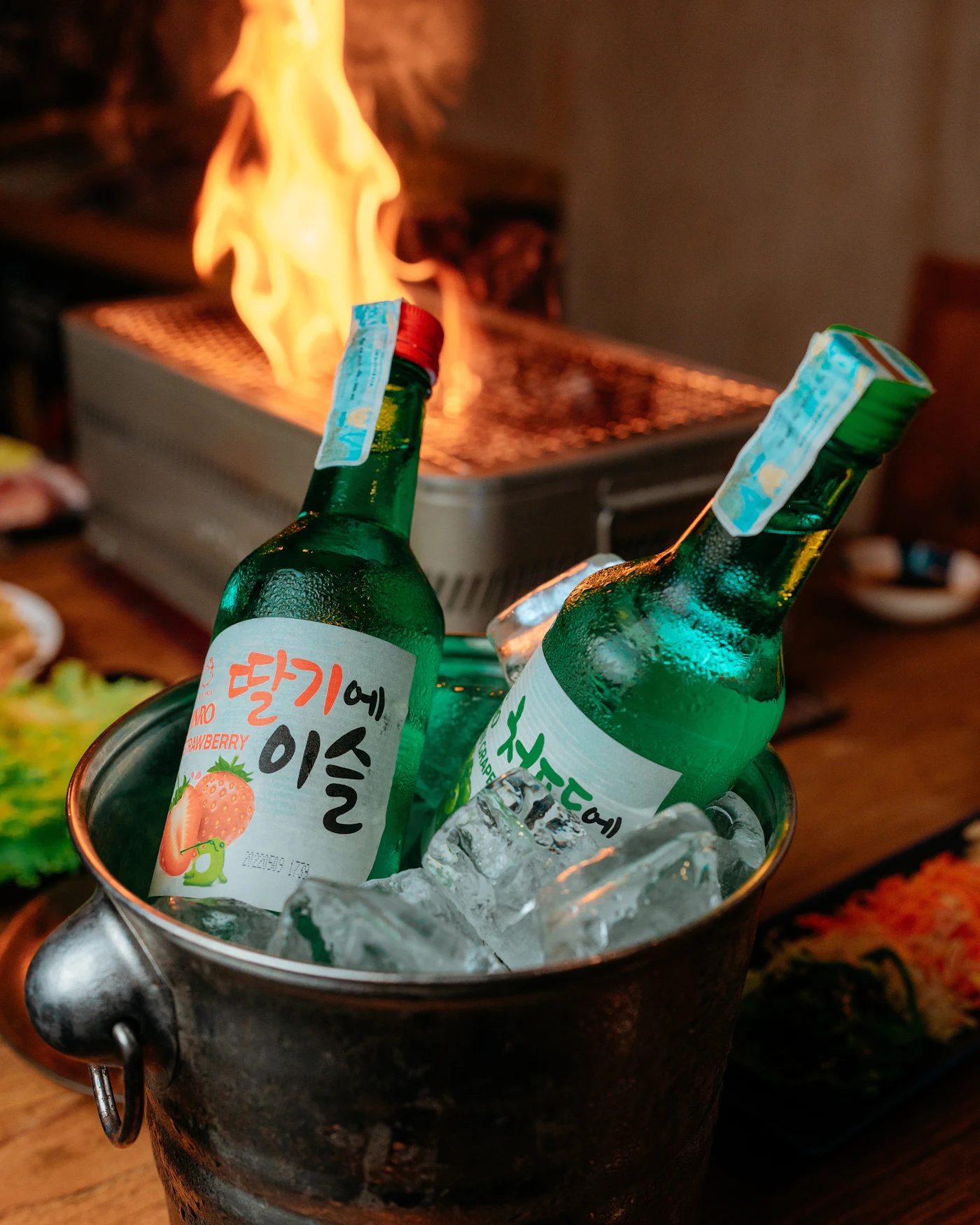 Two green bottles of soju in a bucket of ice in front of a glowing charcoal grill