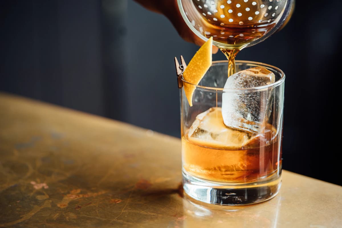 Bourbon being strained over a large ice cube in a rocks glass on the bar