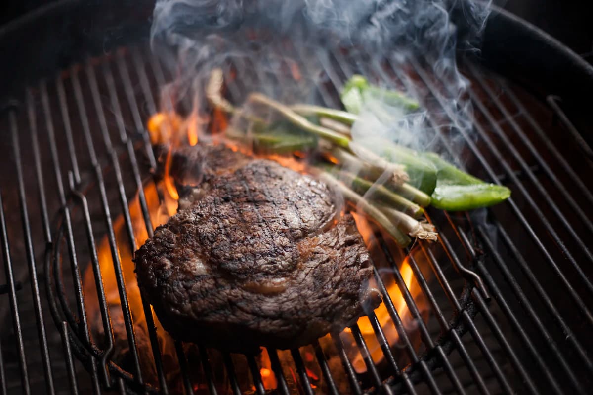 A crusted steak resting on the grill beside charred spring onions
