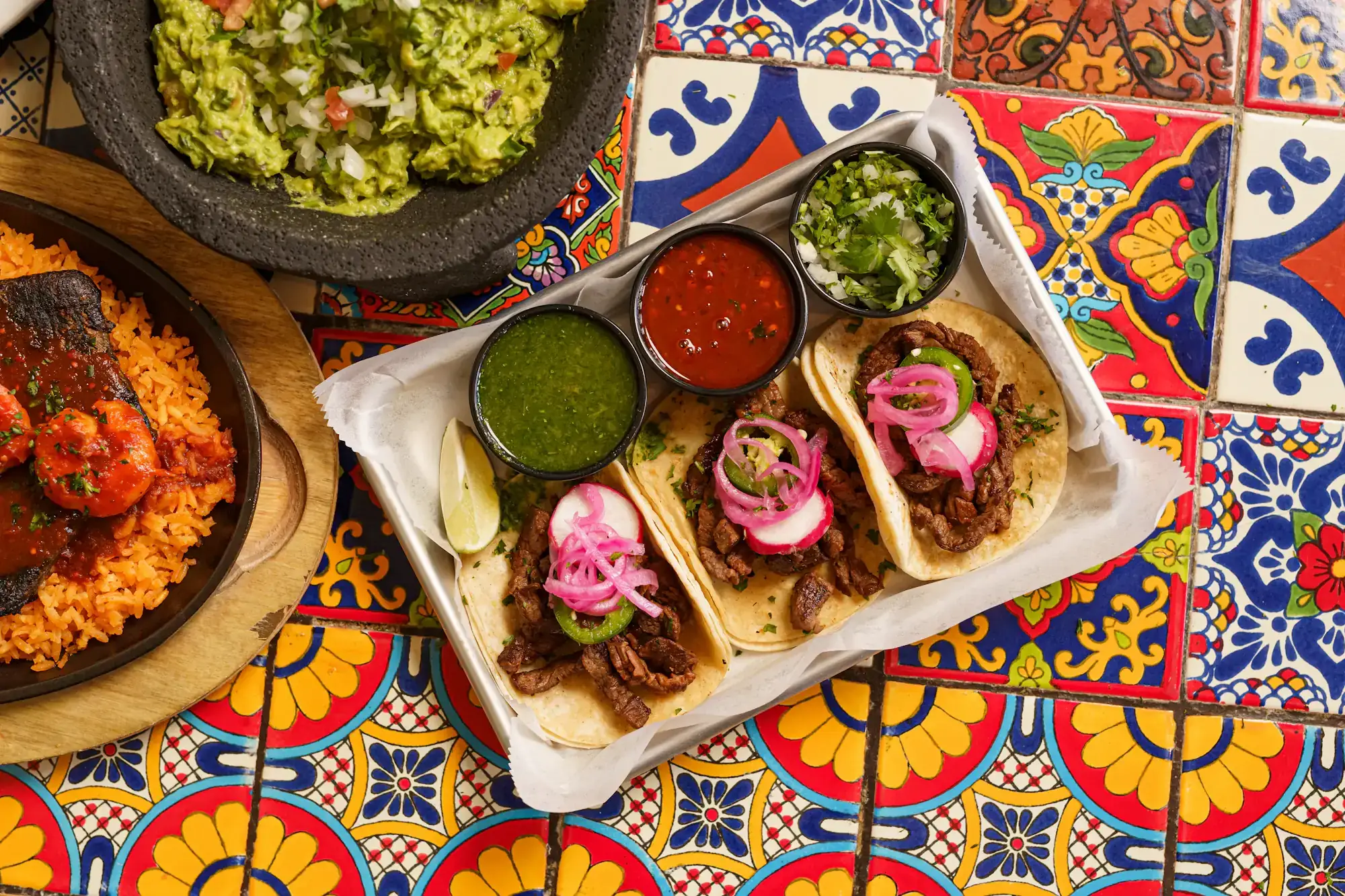 Three tacos with pink pickled onion and three salsas in a paper tray on a painted talavera tile table, guacamole and rice beside them