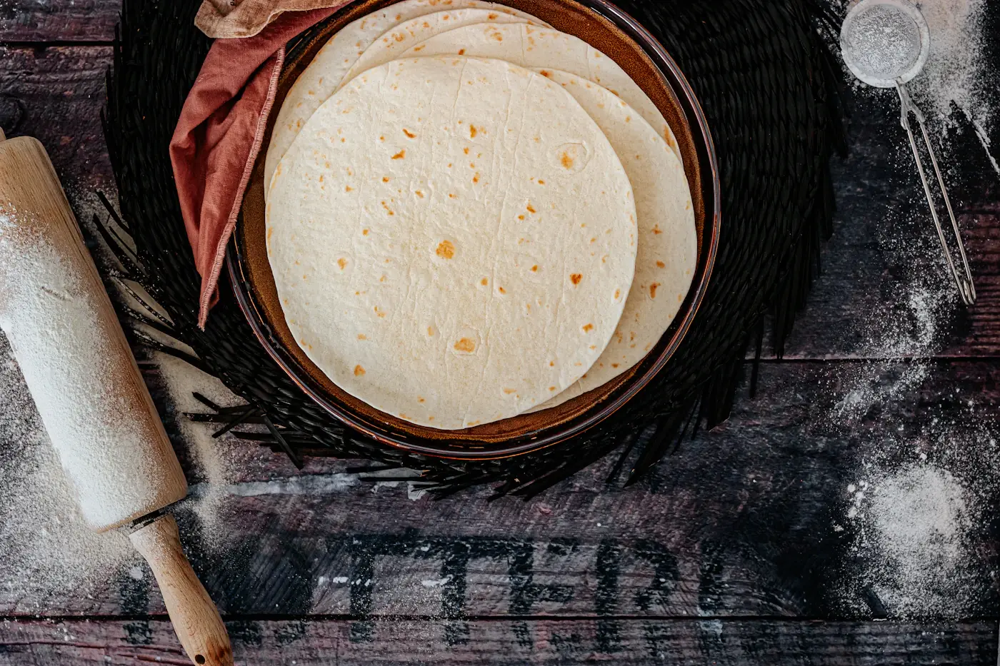 A stack of fresh tortillas in a clay dish on a dark wooden table dusted with flour, a rolling pin beside it