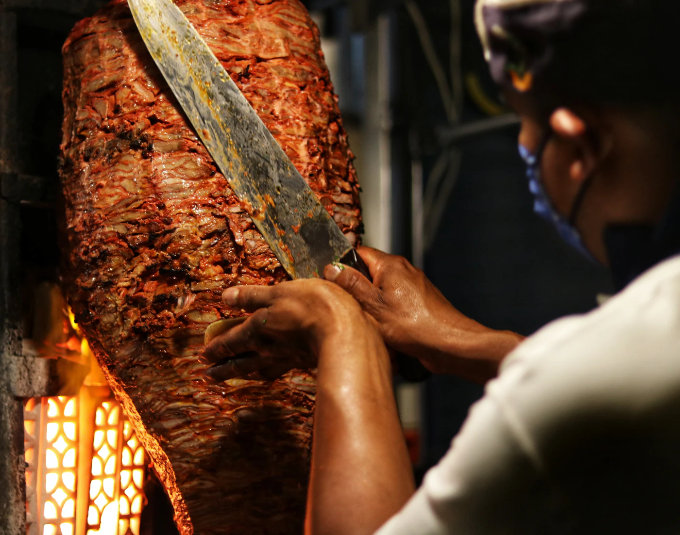 A taquero carving al pastor pork from the vertical spit, the fire glowing below it