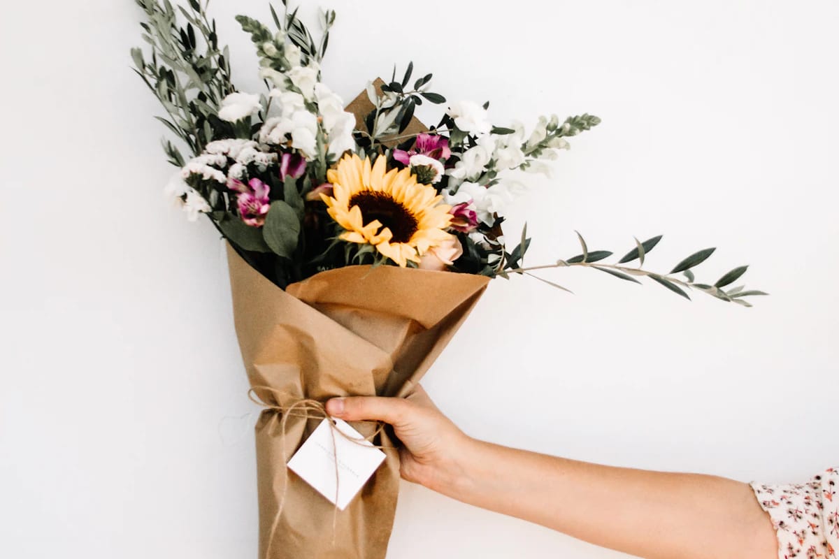 A hand holding out a paper-wrapped bouquet with a sunflower and a gift tag