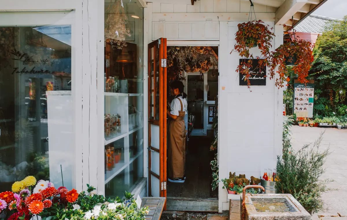 A florist in an apron standing in the doorway of a white flower shop, buckets of flowers outside