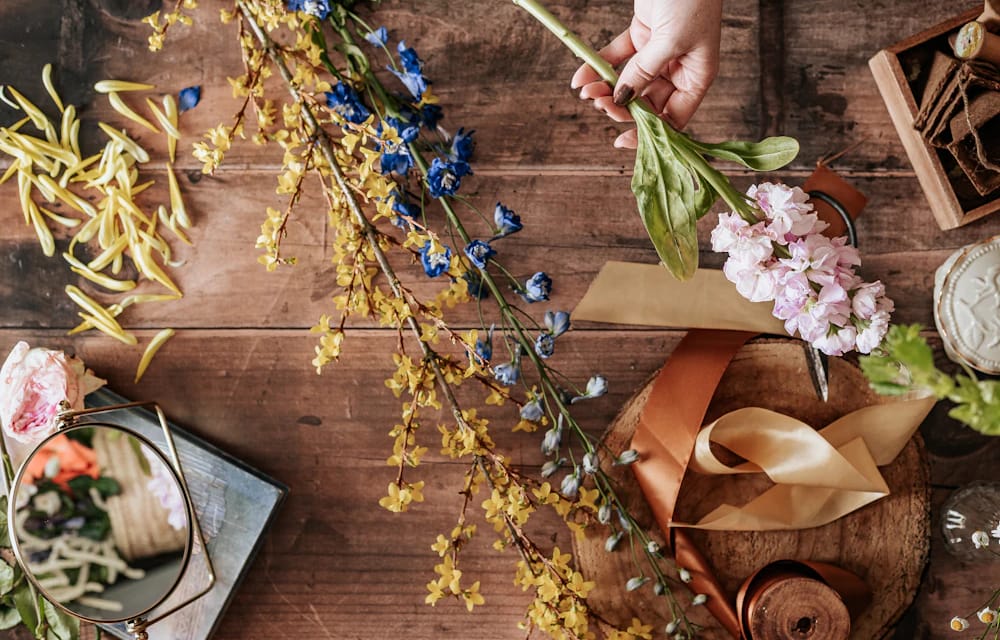 A florist's workbench from above: cut stems, ribbon, scissors and a hand tying a small bunch