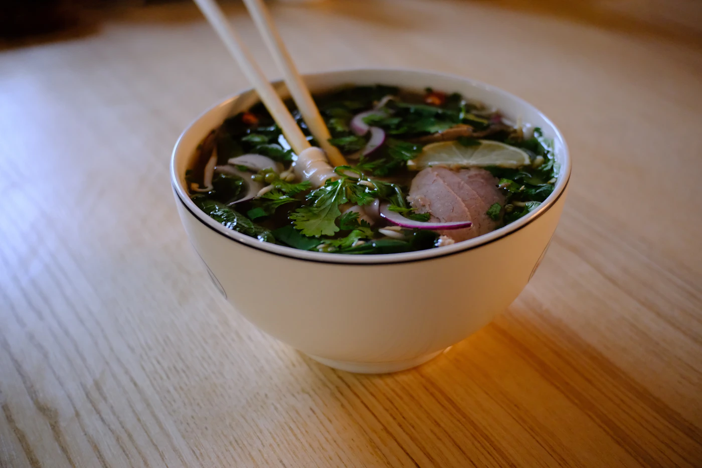 Tô phở bò với rau mùi, hành tây và lát chanh trên bàn gỗ sáng màu, đôi đũa đặt trong bát
