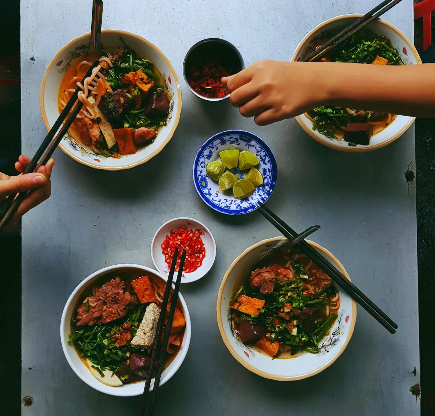 Nhìn từ trên xuống: bốn bát bún, đĩa chanh và chén ớt trên bàn, những đôi đũa đang gắp