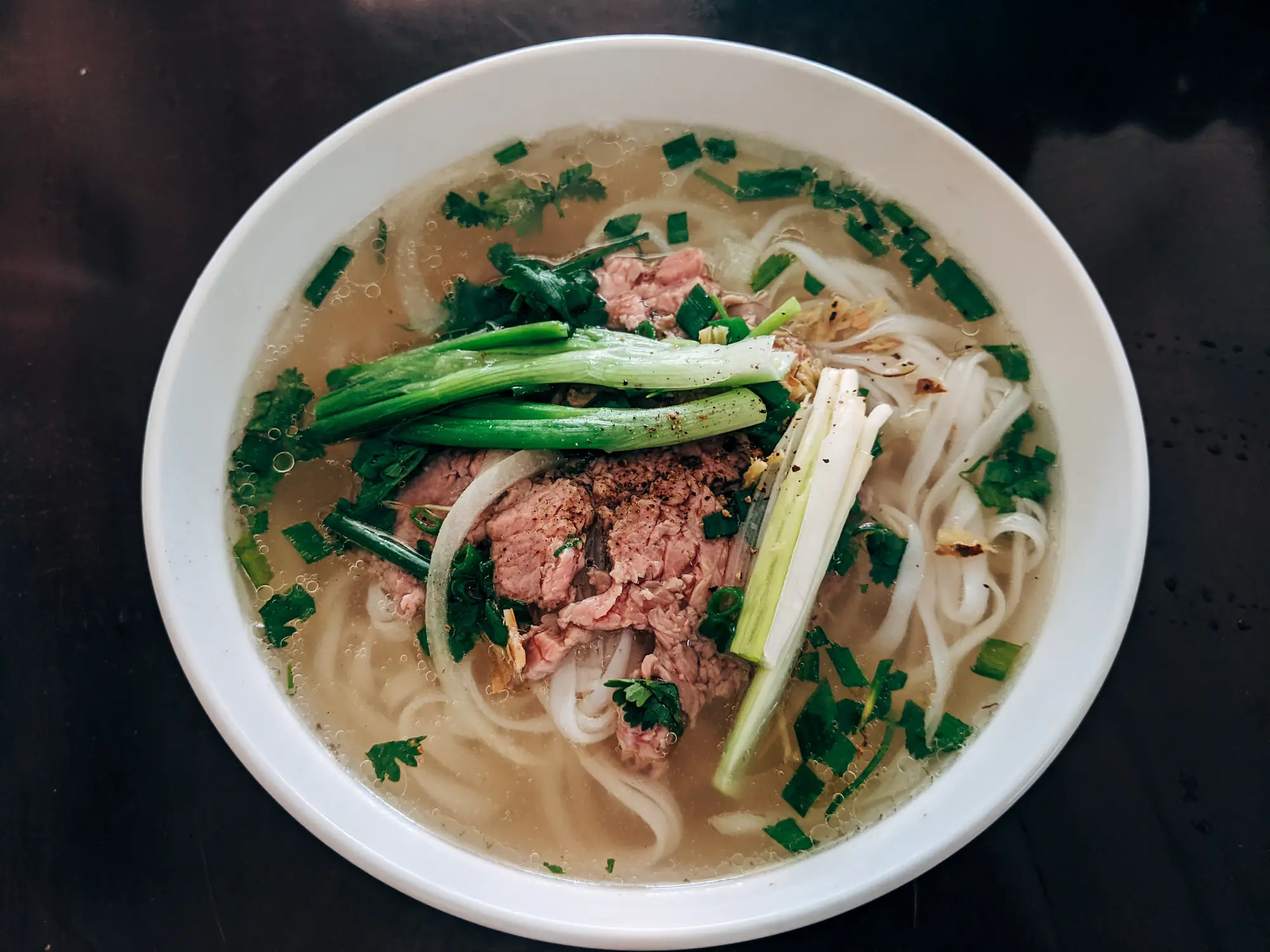 Tô phở bò tái với hành lá, rau mùi và nước dùng trong trên mặt bàn tối màu