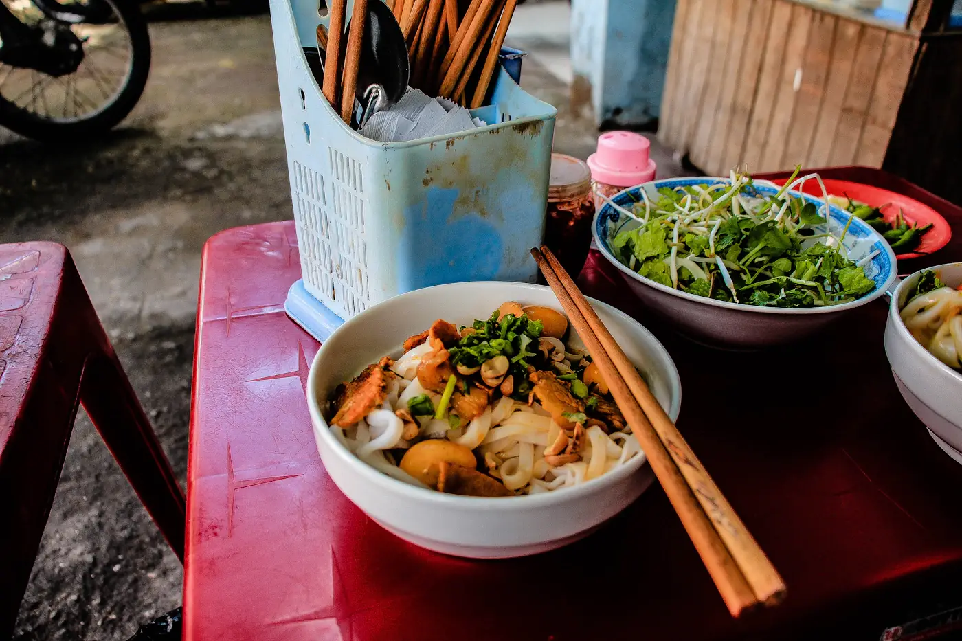 Bát bún nước trên chiếc bàn nhựa đỏ ở vỉa hè, cạnh hộp đũa và bát rau sống