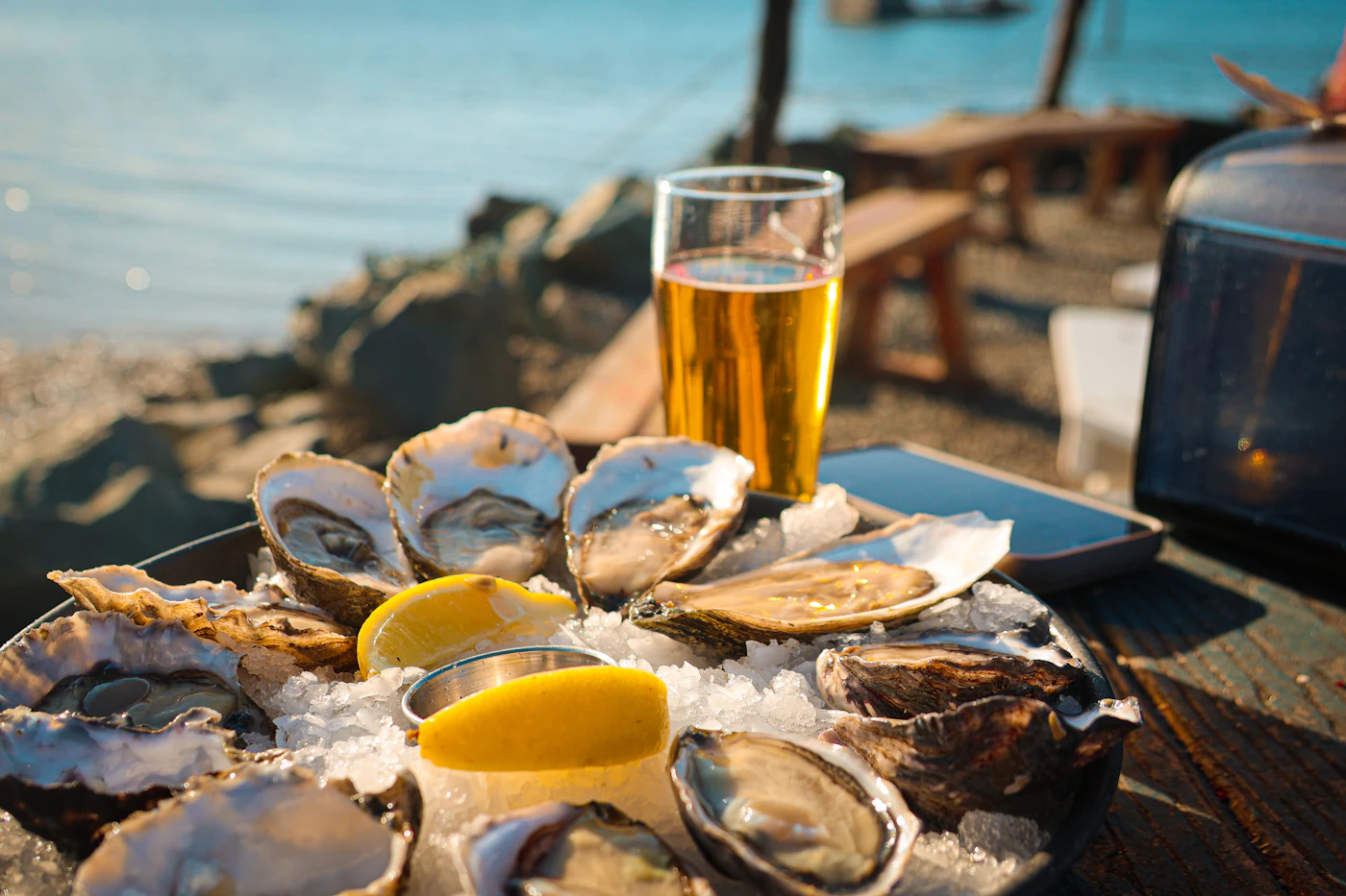 A tray of oysters on ice with lemon next to a glass of beer, on a table by the sea