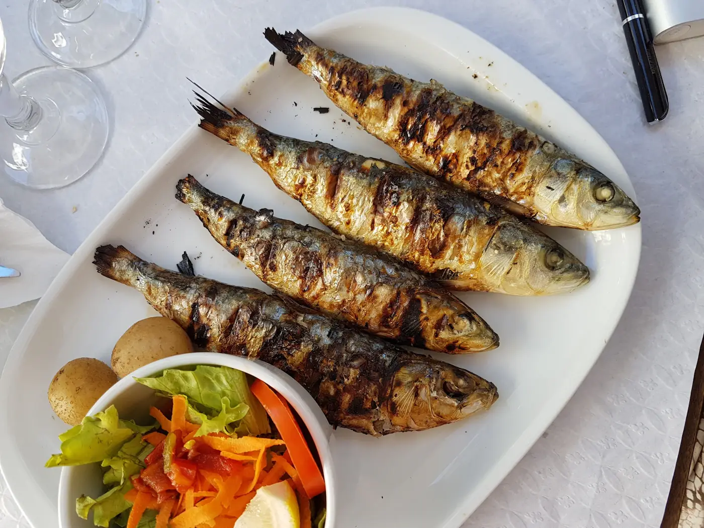 Four grilled sardines on a white platter beside boiled potatoes and a green salad