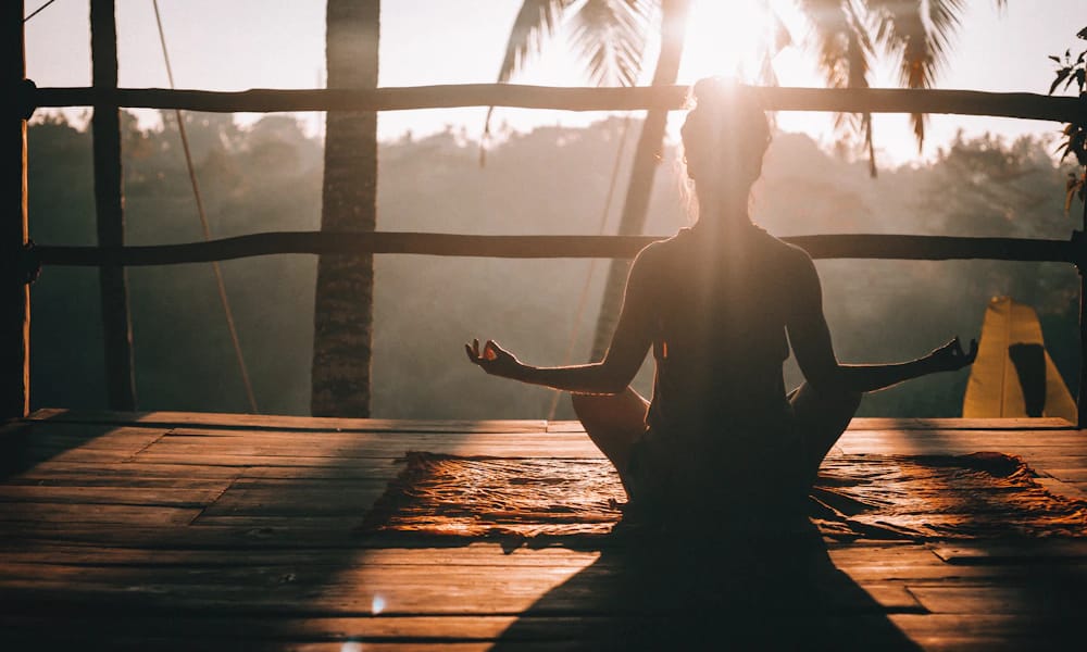 A woman sitting cross-legged on a wooden deck at sunrise, in silhouette against the light