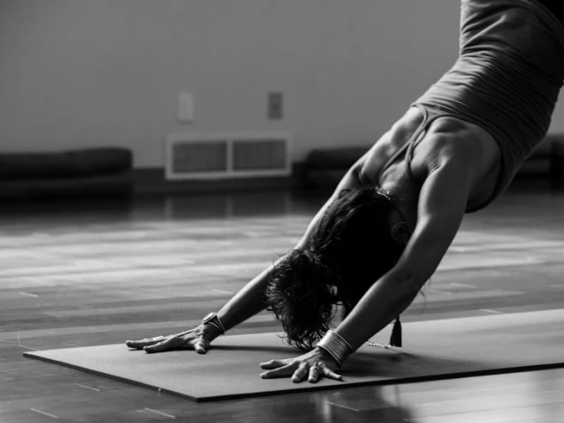 A woman in a downward-facing dog on a mat in a dim, empty studio, in black and white