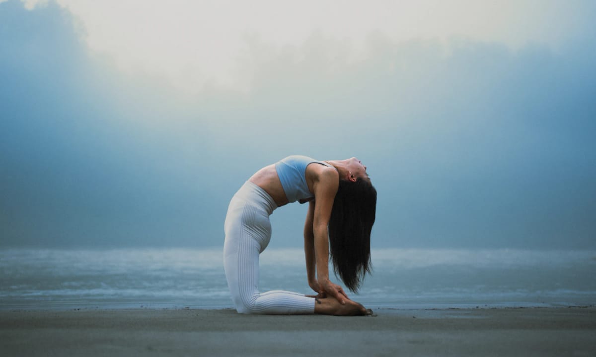 A woman in a deep backbend on a misty beach, the sea flat and blue behind her