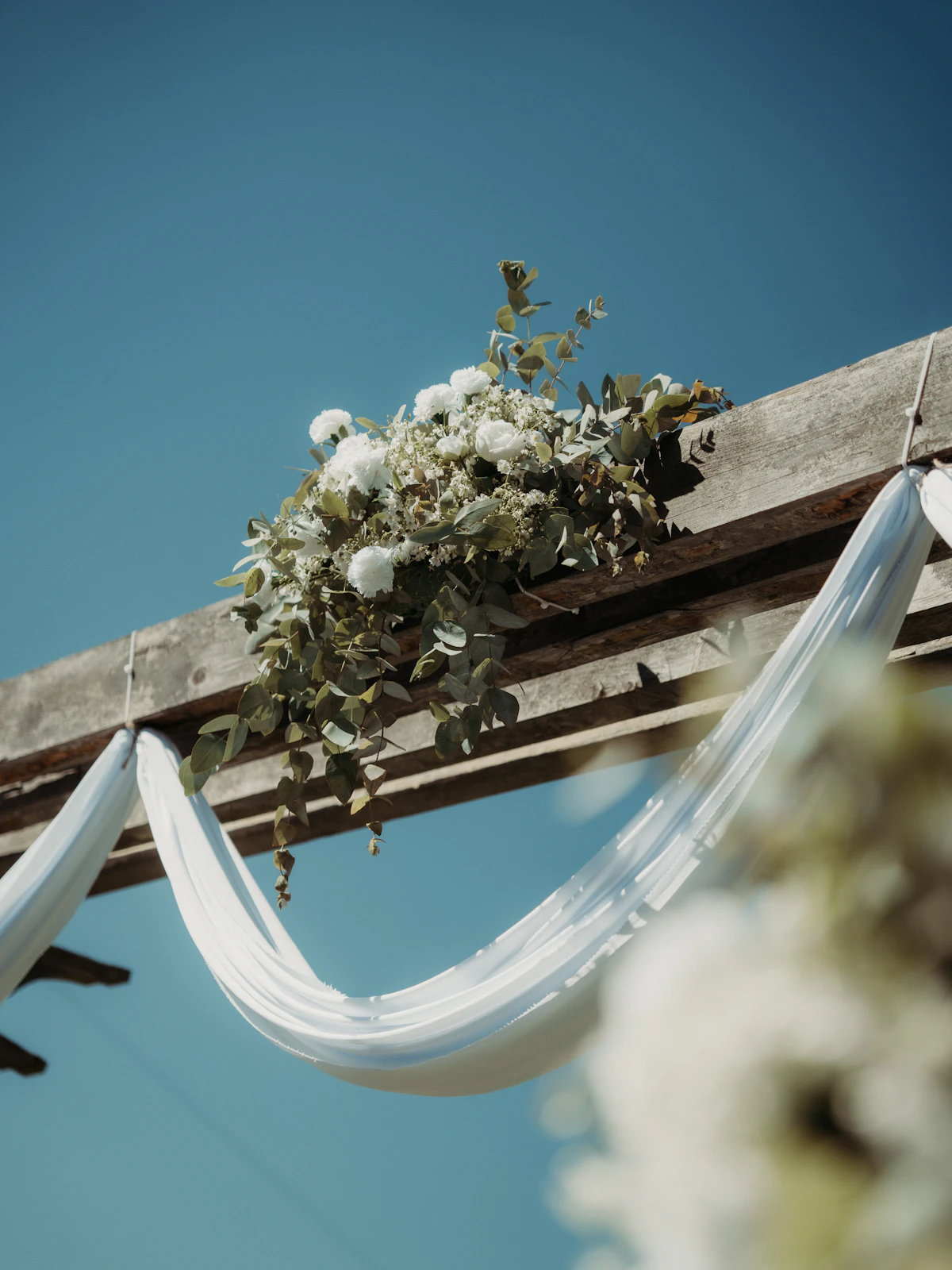 White flowers and greenery on a wooden arbour hung with flowing white fabric
