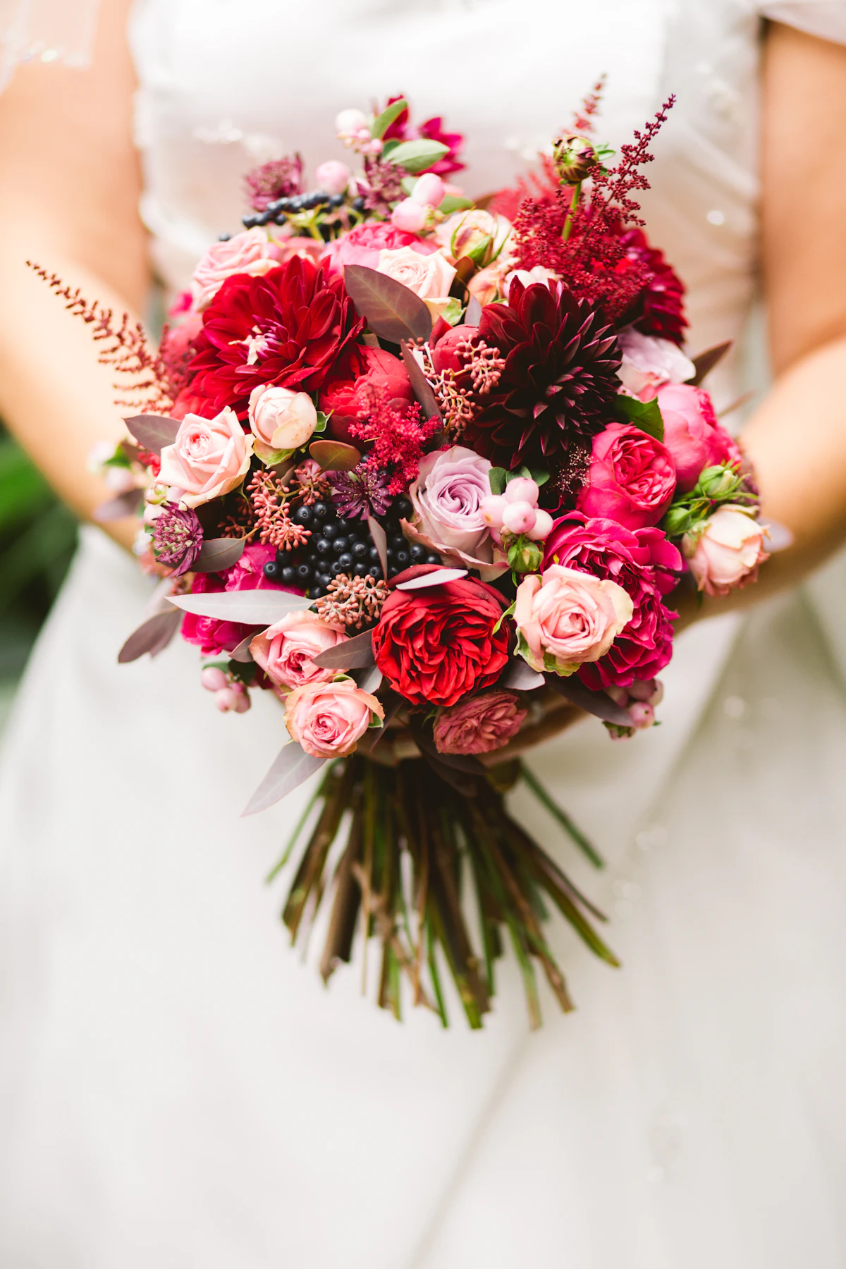 A bride holding a bouquet of red, burgundy and blush roses with dark berries